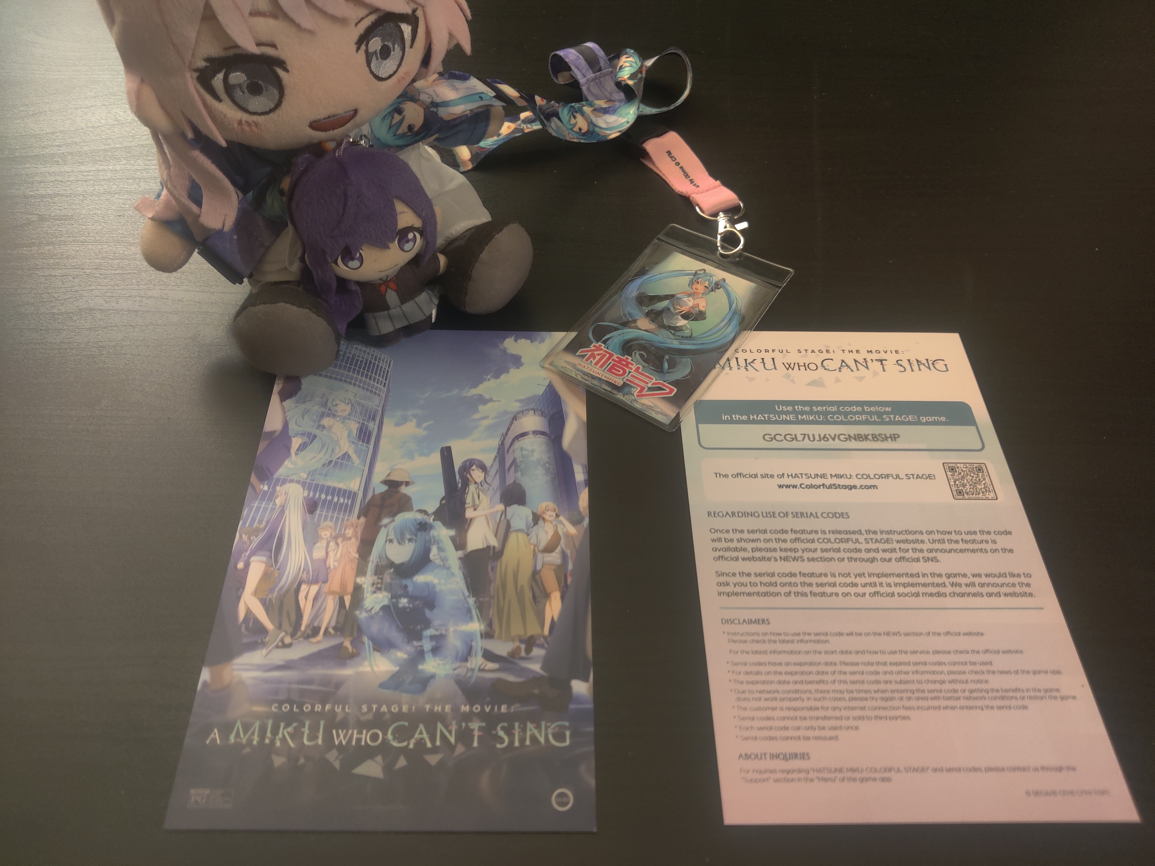 Anon plush and tiny Mafuyu sitting between her legs look at the special miku movie postcards (two of them, one facing up one facing down) and Anon is wearing Miku card lanyard